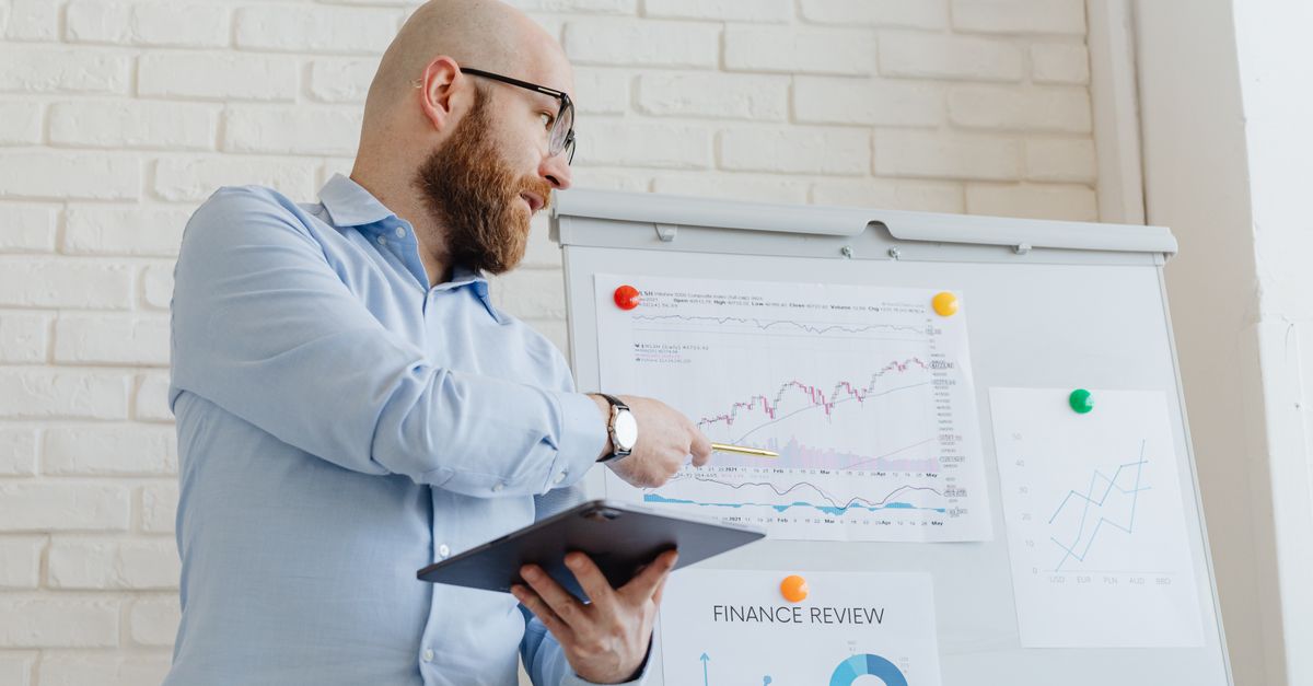 Man giving a business presentation with financial graphs on a whiteboard using a tablet for explanation.