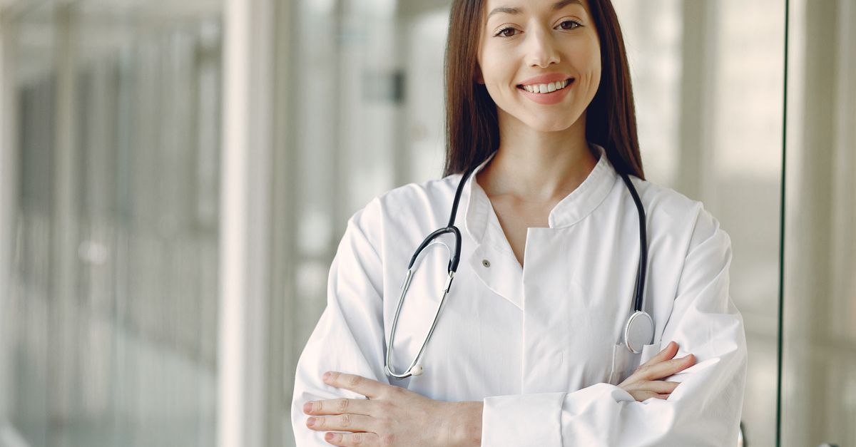 Crop cheerful ethnic female doctor in medical uniform and stethoscope holding hands crossed standing in modern clinic corridor with glass wall