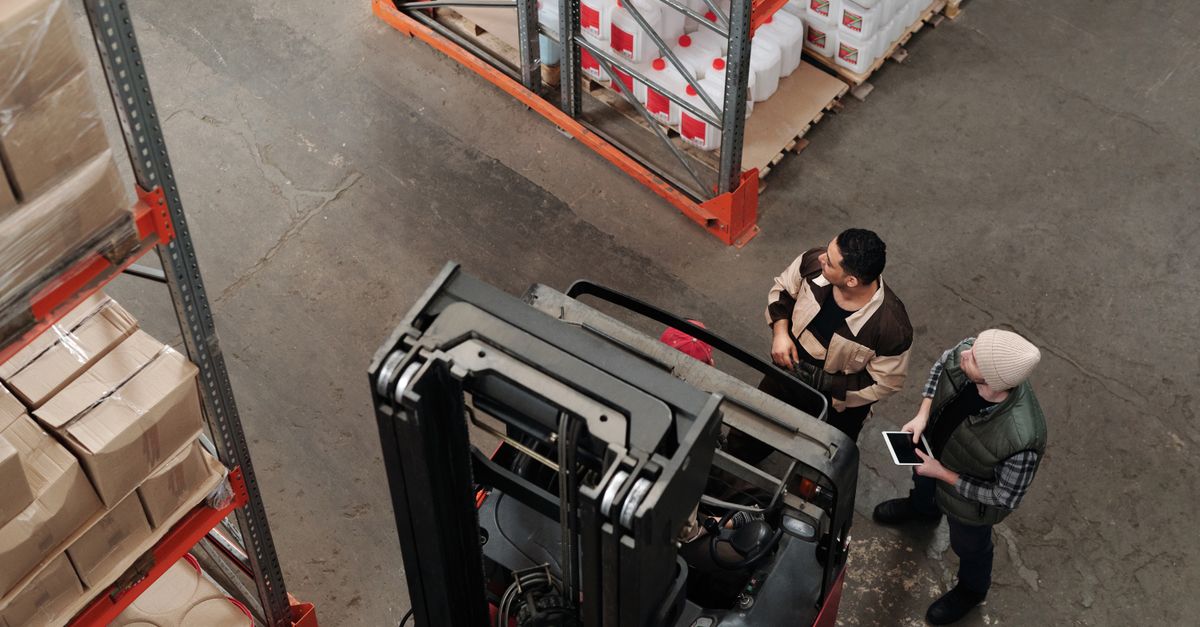 Two workers in a warehouse discussing logistics near a forklift captured from above.