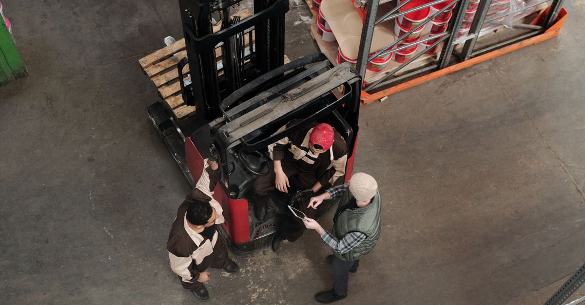 Top view of workers discussing plans in a warehouse beside a forklift, emphasizing teamwork.