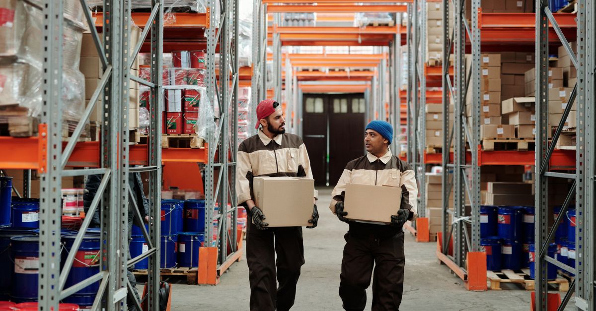 Two workers carrying boxes in a warehouse aisle, engaged in logistics